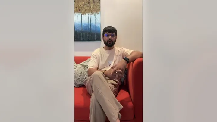 Video still: a guest seated on the red sofa in the lounge, speaking to the camera