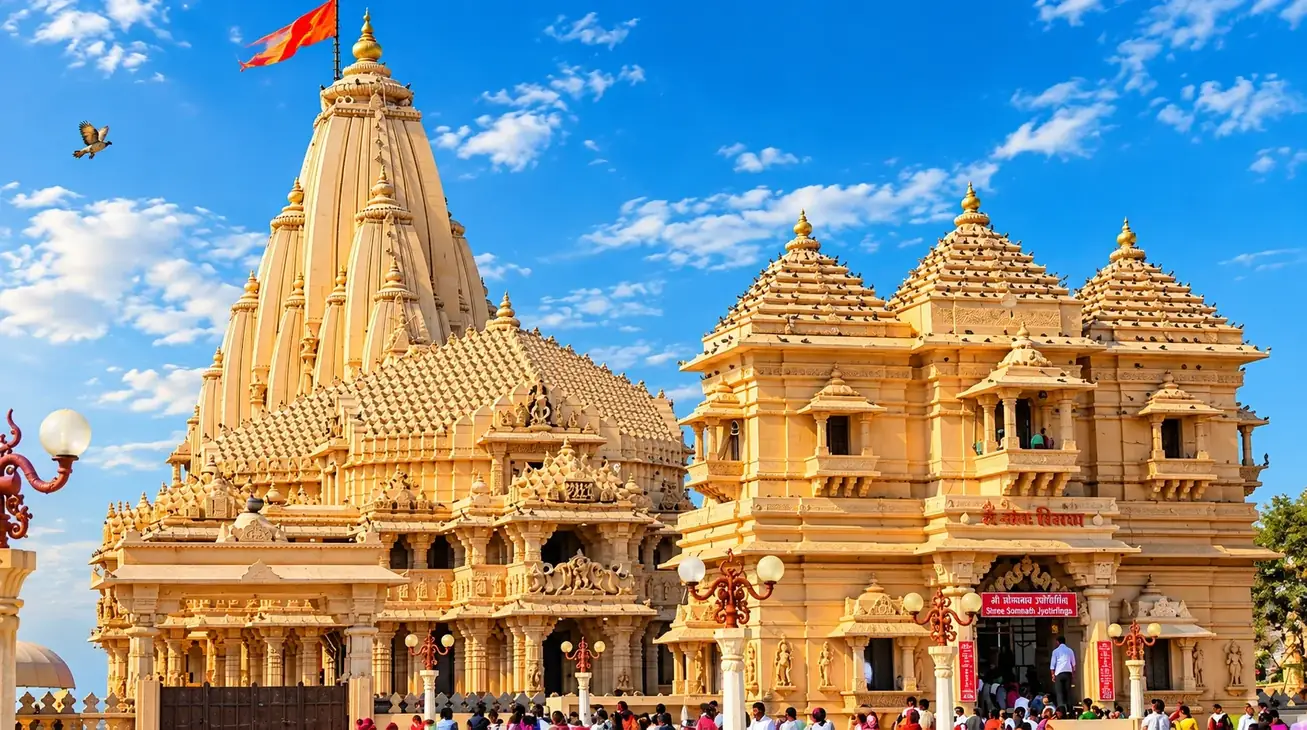 A cream sandstone temple under a blue sky: a tall ribbed shikhara flying a saffron flag beside a pillared hall with pyramidal roofs, ornate lamp posts and a crowd of pilgrims at the gate